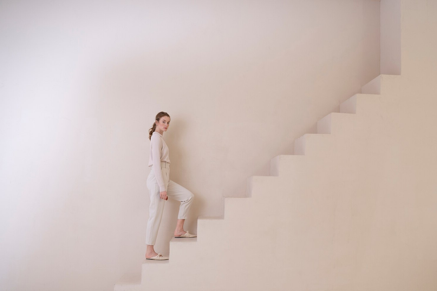 밝은 색 옷을 입은 사람이 미니멀한 계단 위에 서서 올라가는 모습. 환경은 깨끗하고 현대적이며, 부드럽고 중립적인 색조로 차분하고 평온한 분위기를 자아낸다.