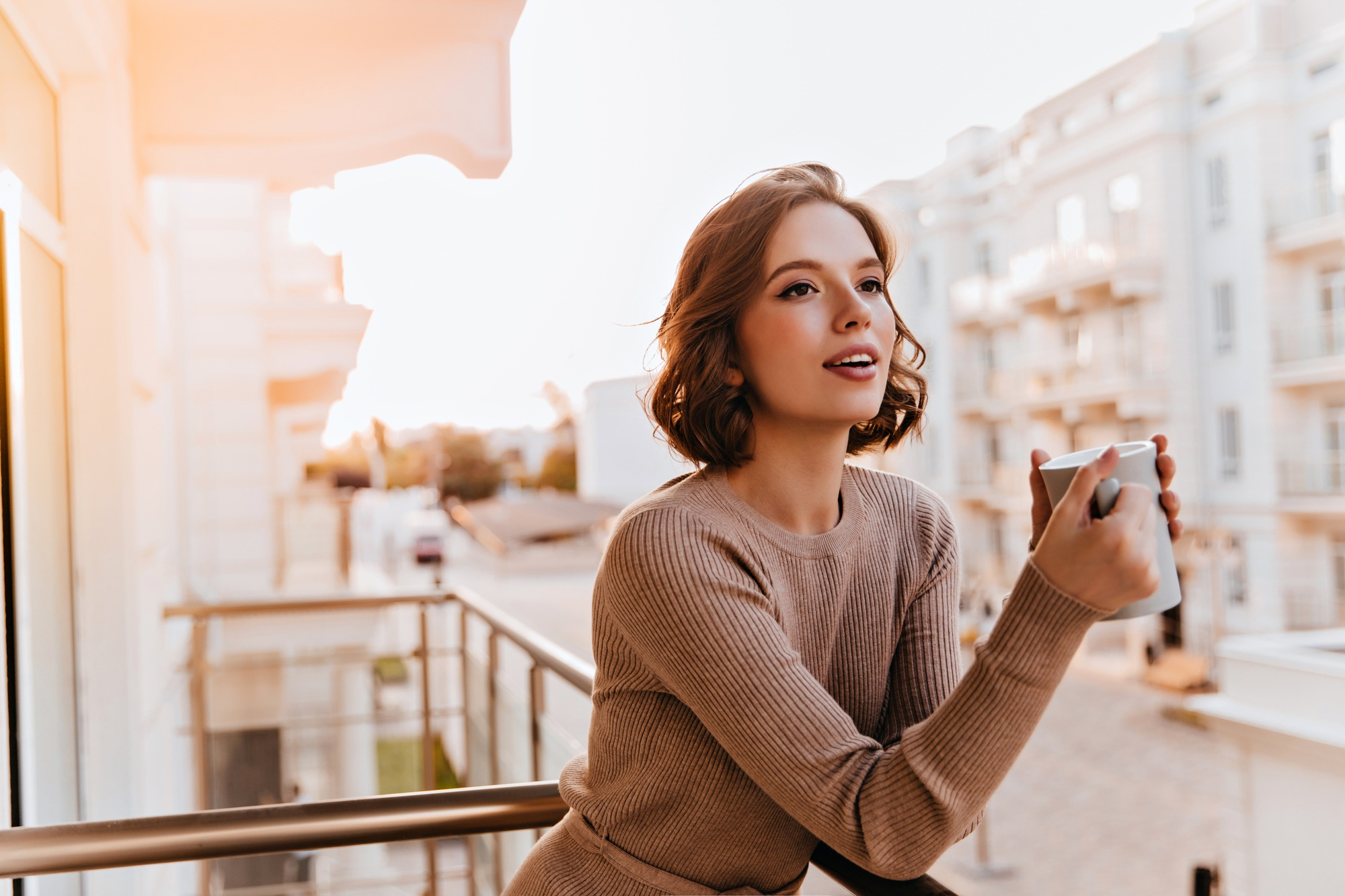 A woman with short brown hair wearing a ribbed beige sweater stands on a balcony, eyes closed and inhaling from a white mug as warm sunlight glows behind her.