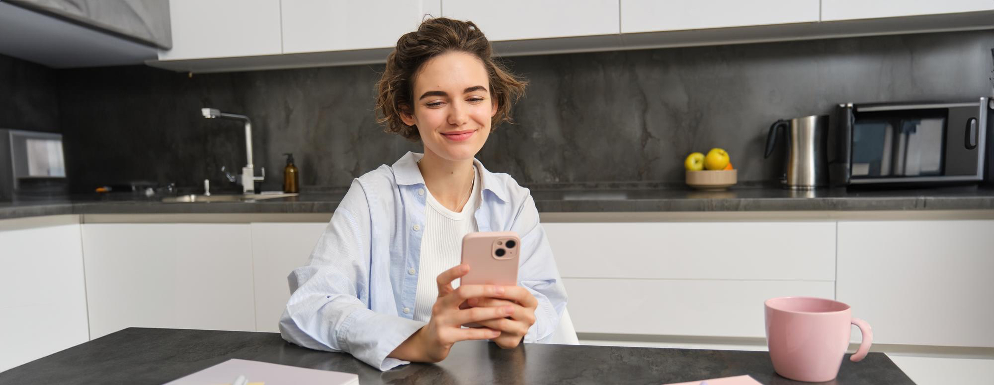 A young woman sits at a kitchen table, smiling as she uses a smartphone to enter her sleep details from last night into the Aeveos app; a pink mug, notebook, and modern kitchen background create a calm morning routine scene.