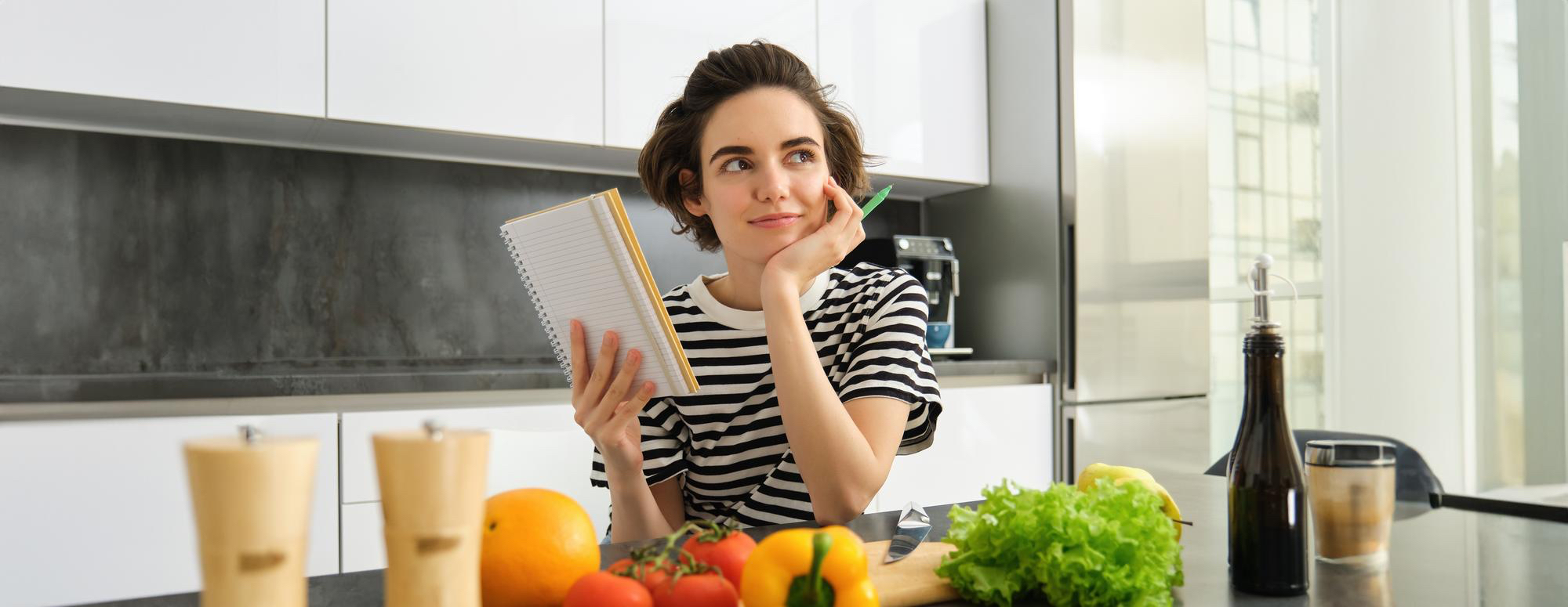 A young woman sits at a kitchen table, smiling as she checks the Aeveos app on her smartphone which displays a personalized "daily plan" for how to live today to sleep better tonight; a pink mug and notebook sit nearby on the table, and the bright, modern kitchen in soft morning light creates a calm, focused routine atmosphere.