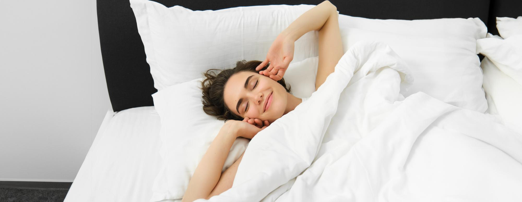 A young woman lies in bed with a peaceful, satisfied expression, waking up refreshed after following the Aeveos app's personalized daily plan to improve her sleep; soft white bedding and morning light emphasize a calm, restorative morning.