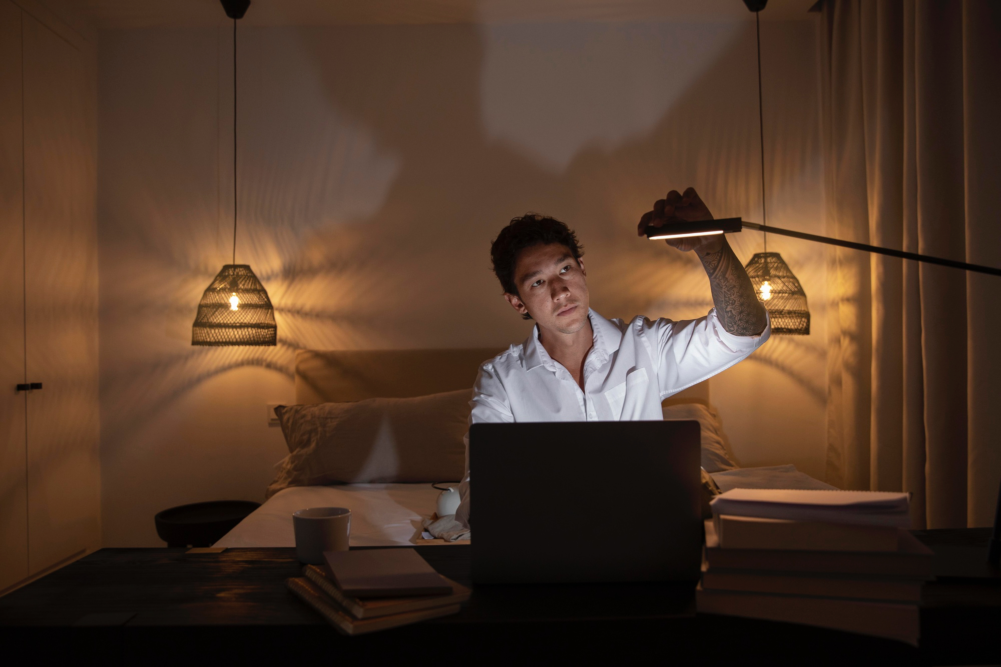 A man adjusts a bedside lamp while checking the Aeveos app on his phone, following the app's sleep plan to dim and tune the room lighting for better sleep. The bedroom is softly lit with warm lamps and a laptop on the desk, creating a calm, pre-sleep atmosphere.