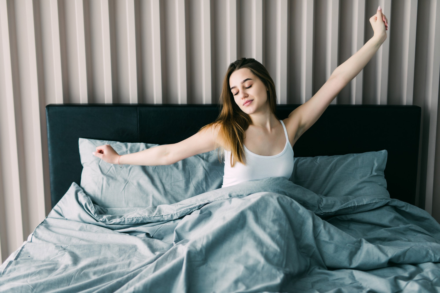 A woman sits upright in bed stretching both arms overhead, wearing a white tank top. The bed is covered with gray sheets and pillows, set against a dark upholstered headboard and a wall with vertical beige panels. Soft daylight fills the room, creating a calm and refreshed morning atmosphere. The emotional tone is peaceful and energizing, suggesting the start of a new day.