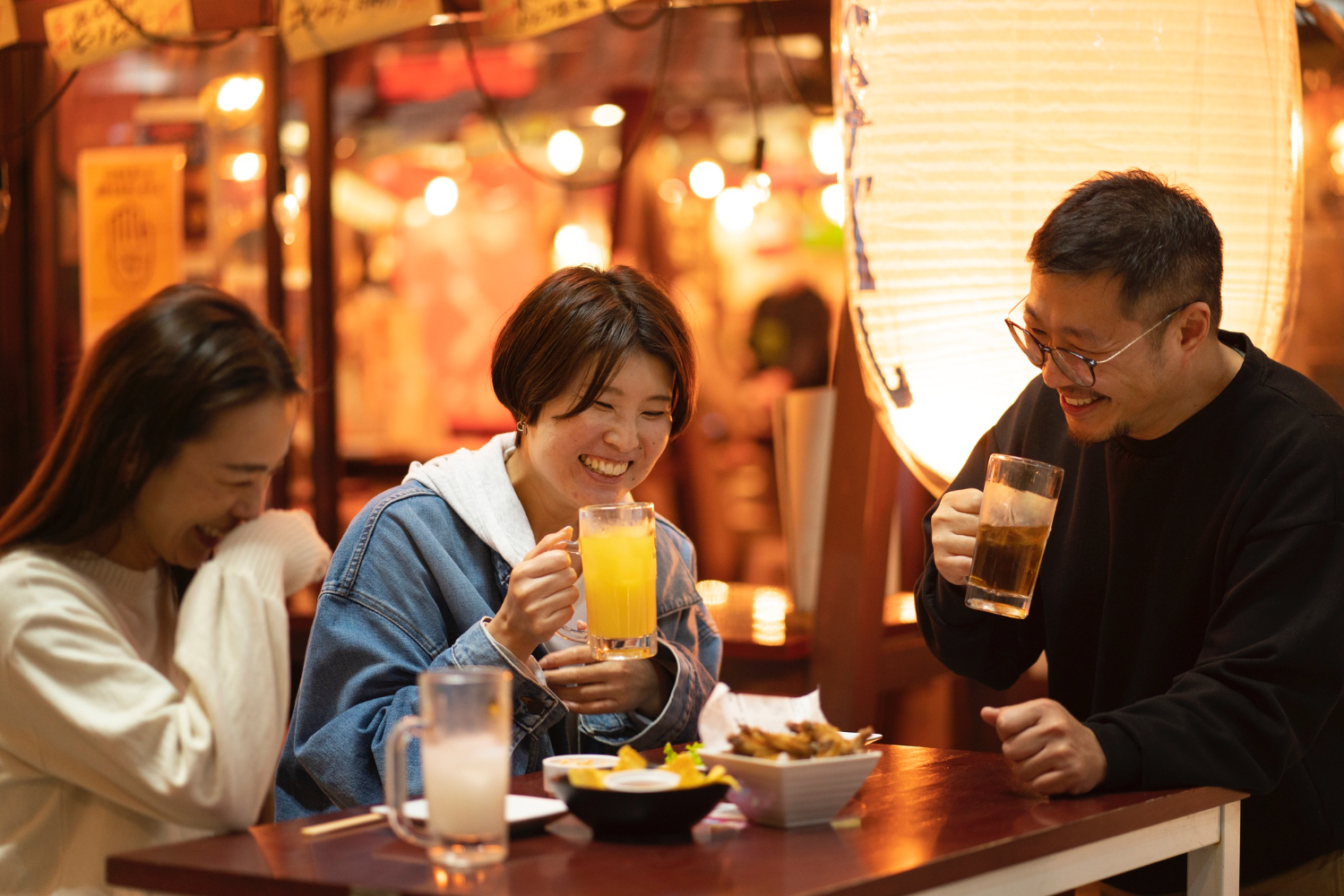 Three people sit together at a wooden table in a warmly lit restaurant, each holding a glass of beverage and engaging in conversation. The table is set with plates of food and drinks, and the background features decorative lights and a large paper lantern, creating a cozy and lively atmosphere. The mood feels relaxed and social, suggesting friends enjoying a meal out.